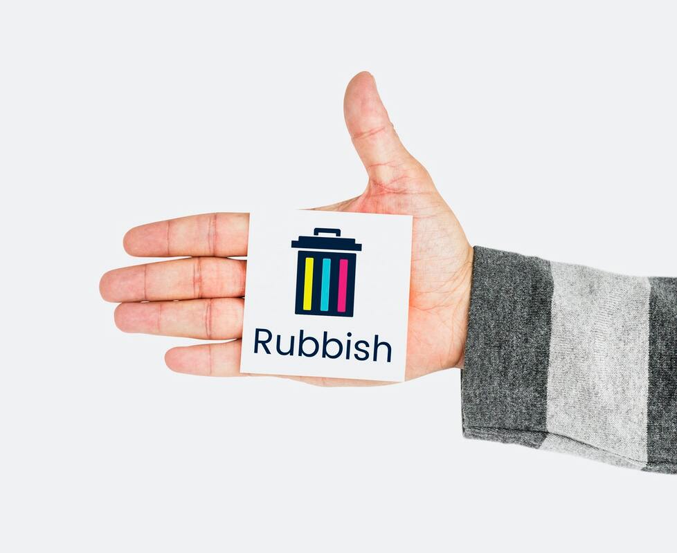 A right hand with the palm facing the viewer and the thumb pointing up. The palm is open and stuck to it is a white post-it note with a picture of a black, yellow, blue and pink waste bin. Under the bin is the word rubbish. 