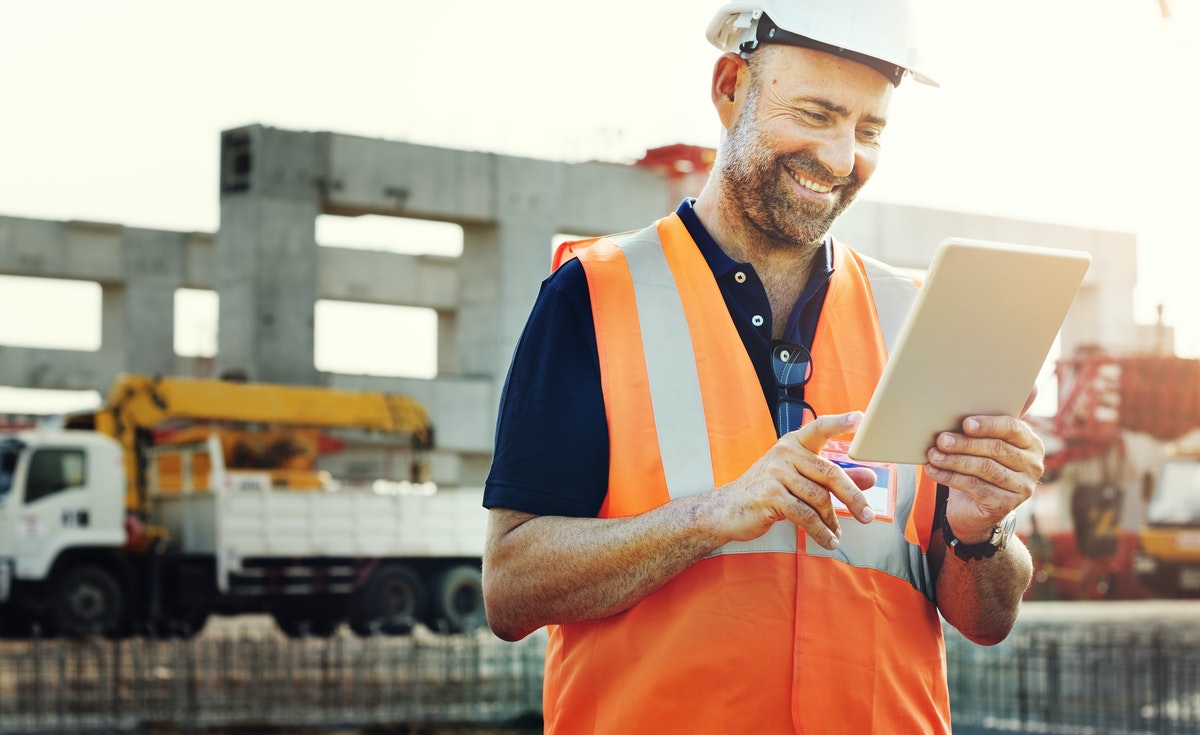 A man is on a building site. He is smiling and holding a tablet computer. He is wearing a dark blue polo shirt with glasses hanging from the buttons. He is wearing a hard hat and an orange high visibility vest. Construction work is depicted in the background.